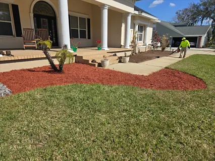 residential landscape with silage tarp garden bed (red mulch), worker using leaf blower near columned house porch