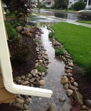 residential rain gutter garden: white downspout directs rainwater into a decorative stone-lined stream with ornamental plants, surrounded by mulch beds and lawn during rainfall