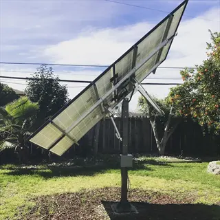 residential solar tracker garden with dual-axis photovoltaic panel array mounted on steel pole in sunny backyard; surrounded by citrus tree, palm plants and wooden fence