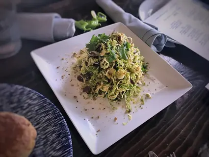 square white plate of shredded brussels sprout salad with nuts, herbs, and cheese, served on a dark wooden table with menu text and other dishes