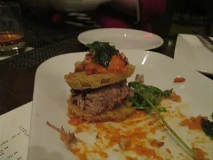 stacked stuffed green tomatoes with sauce and arugula on a white plate in a restaurant setting, with menu text visible in the background