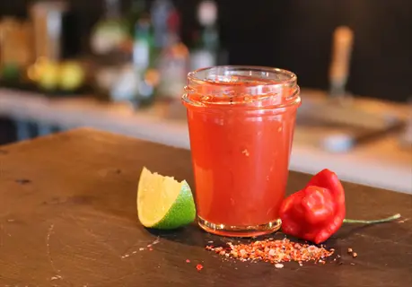 tomato cocktail drink in a glass with lime wedge, red chili pepper, and seasoning on a wooden bar counter, bar background with bottles