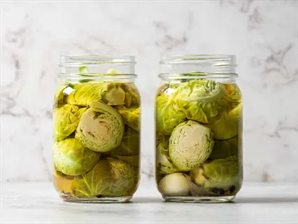 two glass jars of fermented brussels sprouts preserved in brine, set against a light marble background