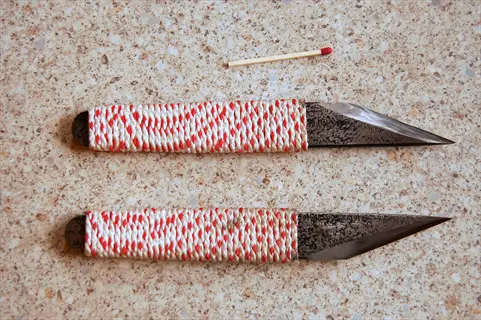 two grafting knives with red-and-white wrapped handles on textured surface, matchstick for scale