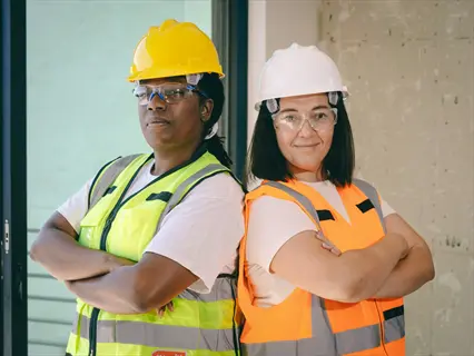 two workers in high-visibility vests and hard hats wearing safety gardening glasses stand back-to-back indoors