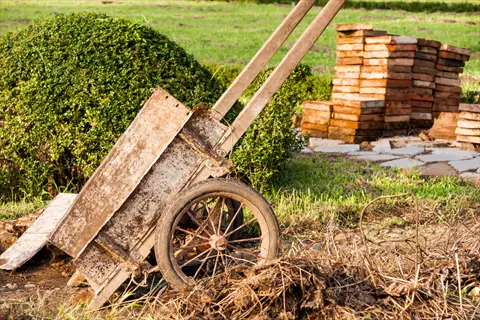 weathered wooden hand truck in garden setting with trimmed bush and stacked bricks. (hand truck garden context: used for transporting materials in outdoor cultivation projects)