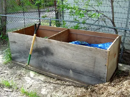 wooden backyard compost bin with pitchfork and blue tarp in a fenced garden area surrounded by soil and vegetation