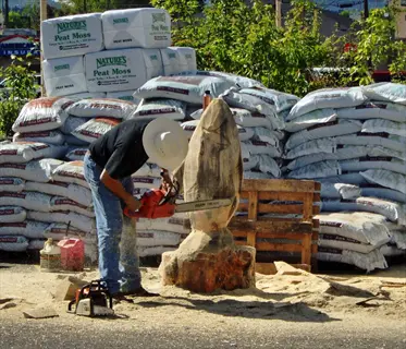 worker chainsawing a log near stacked peat moss bales labeled 'nature's peat moss' in an outdoor setting