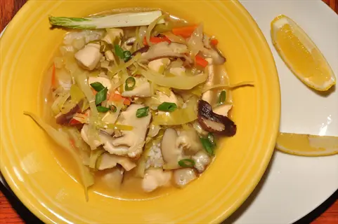 yellow bowl of vegetable stir fry with lemongrass, chicken, mushrooms, cabbage, and carrots, garnished with green onions and lemon slices