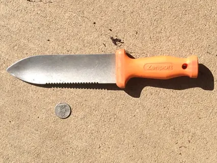 zenport hori hori garden knife with orange handle and serrated stainless steel blade on concrete surface, shown with quarter for scale